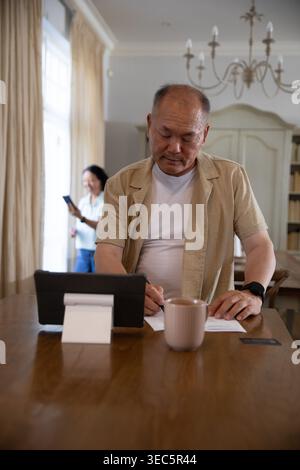 Asian couple, senior woman using smartphone near window, man signing papers using tablet at table Stock Photo