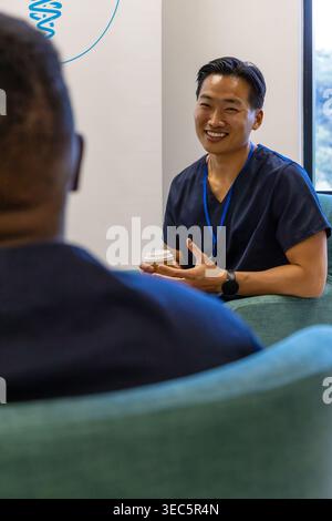 Diverse male coworkers chatting in clinic lounge, navy scrubs with blue lanyard holding coffee cup Stock Photo