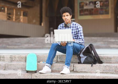 Young Man Using Laptop While Sitting on Steps With Backpack and Skateboard Nearby Stock Photo
