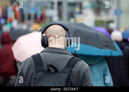 Man in headphones walking on background of people with umbrellas, rain in city Stock Photo