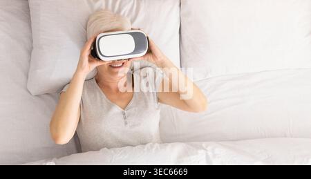 Senior woman lying on bed and using virtual reality headset Stock Photo