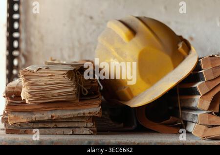 Worn yellow helmet on a rusty metal shelf, leaning against stacks of handwritten ledger books from the late 1980s in an abandoned space. Stock Photo
