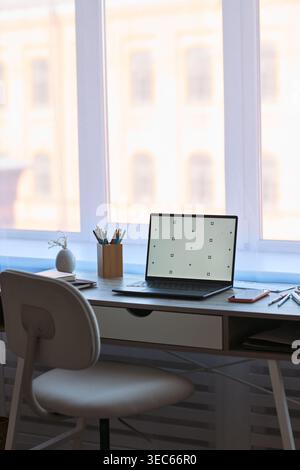 Vertical shot of home office desk featuring laptop with blank screen in spacious room  Stock Photo