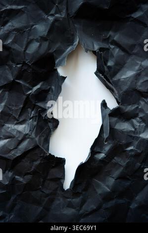 Torn black crumpled paper with central hole revealing white background, abstract texture symbolizing damage, break or reveal. Stock Photo