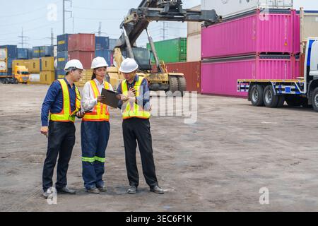 A team of three Asian workers in safety gear review data at a busy Thailand port with heavy machinery. Represents industrial operations, equipment man Stock Photo