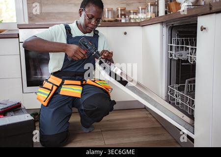 Female technician using electric screwdriver while repairing dishwasher in a kitchen Stock Photo