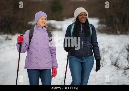 Two diverse women friends enjoying winter hiking on a snowy trail with trekking poles Stock Photo