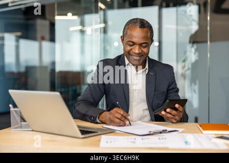 A professional mature African American man, possibly an accountant or financial analyst, using a calculator and writing on documents at his office des Stock Photo