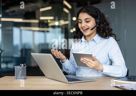 Happy female customer support specialist with headset using laptop and digital tablet in modern office. Online consultant, remote operator, and virtua Stock Photo