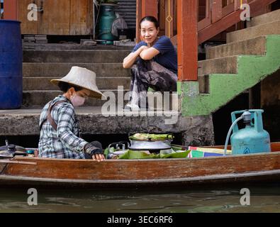 Floating food vendor cooks from a wooden boat as a woman watches from steps at Damnoen Saduak Floating Market in Thailand Stock Photo