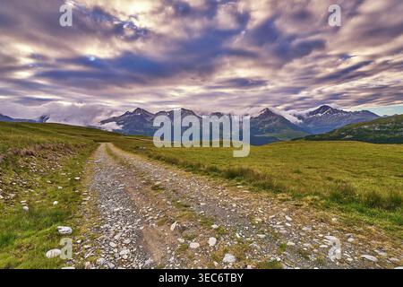Mountain dirt road leading through alpine meadow toward dramatic cloudy peaks in Valchiavenna, Italian Alps, scenic landscape Stock Photo