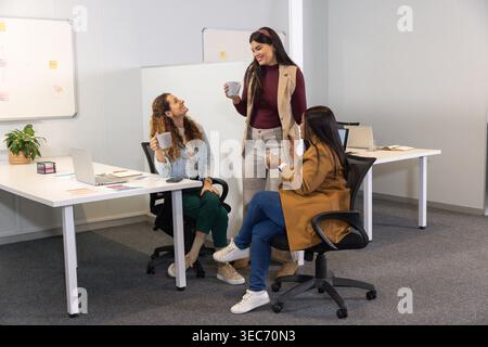 Three female coworkers chatting while holding coffee mugs at modern office desks with laptops Stock Photo