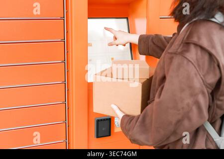 Woman using an automated orange parcel locker to send or receive packages. Holding cardboard boxes while using the touchscreen for delivery or pickup. Stock Photo