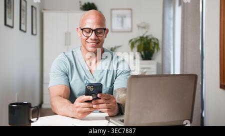Smiling Healthcare Professional Using Smartphone and Laptop for Telemedicine Consultation in Home Office Stock Photo