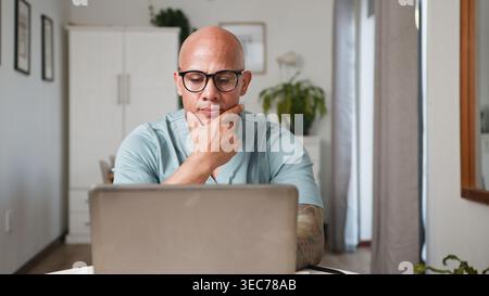 Thoughtful Man Working on Laptop in Modern Home Office Remote Work Environment Stock Photo