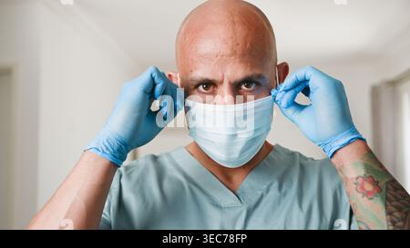 Healthcare Worker Putting On Surgical Mask and Gloves in Clinical Setting With Tattooed Arm Stock Photo