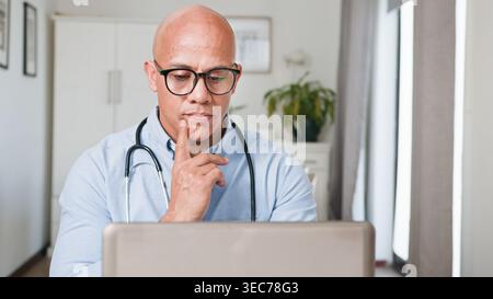 Pensive Male Doctor Working On Laptop In Home Office With Stethoscope For Telemedicine Stock Photo