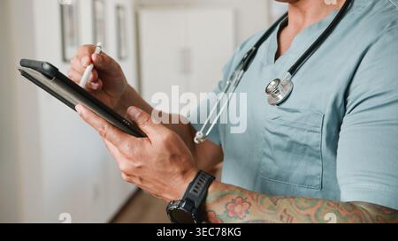 Tattooed Medical Professional in Scrubs Using Tablet With Stethoscope for Digital Charting Stock Photo