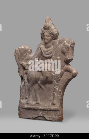 Figurine of Harpocrates-Heron on horseback, Egyptian deities, Harpocrates mythology, Heron mythology, horses. Figurine of a deity sitting on a horse with a wreath and a double crown on his head, dressed in a short military tunic and cloak. The horse is walking to the right, the deity is facing the viewer, to the right. Behind the horse's rump is a palm tree, 2nd century, Egypt, figurine, height 29 cm, width 19.3 cm, The image shows a carved stone relief of a figure seated on an animal, set against a plain gray background; the stone has a weathered, rough texture with visible cracks and chips, Stock Photo