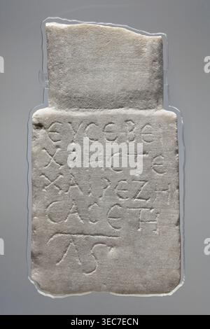 Epitaph of Eusebius, stele in the shape of two rectangular slabs of different widths placed narrower above the wider one. The lower slab has five lines of Greek text. 2nd–3rd century paleography. Sidon. Tombstone inscription in Greek. Height 24.5 cm, width 13.7 cm, depth 3.6 cm., A rectangular stone slab with an irregular top edge resting on a neutral background; the slab is a pale off-white color with subtle gray and beige variations and a slightly rough, weathered surface texture. The front face shows a group of shallow incised linear markings forming repeated shapes across the surface, and Stock Photo