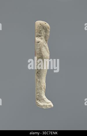 Ushabti, Mummy-shaped figurine, standing on a base with a flat back. Arms crossed on the chest, right folded over left; a pickaxe in the left hand and a hoe in the right. Wearing a smooth, three-part wig with ears uncovered. Facial details illegible; long beard damaged. No inscription. Date: 664–30 BC Late Period–Ptolemaic Period. Place of origin: Memphis, Egypt. Shabti, height 6 cm., A pale beige weathered vertical object with an irregular, elongated rounded top and a slightly protruding lower end resembling a curved foot, showing a rough, pitted surface with small cracks and textured Stock Photo
