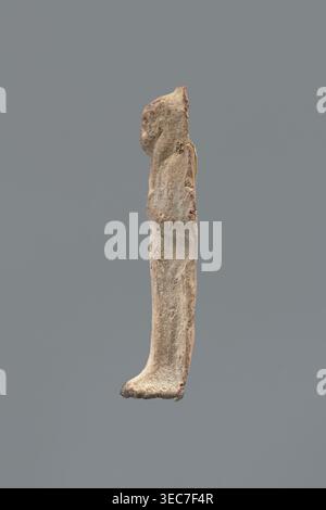 Shabti, Mummy-shaped figurine shabti, funerary gift; made carelessly, impressed in a mold. Hands folded on the chest, right over left; both hands hold hoes. Wears a three-part smooth wig. Facial details poorly legible. No inscription. Back unfinished. c. 1069–c. 656 BC, Third Intermediate Period, Egypt. Height 7.5 cm., A vertical, elongated object resembling a narrow carved figure displayed against a solid gray background, with a rough, weathered surface in shades of beige, tan, light brown, and hints of reddish-brown, showing irregular contours and a rounded top, a slightly curved midsection, Stock Photo