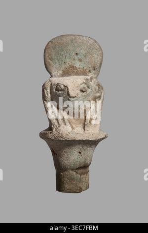 Amulet - falcon head on a column; papyrus capital of the column shows a falcon head in a striped wig topped with a disc, with a pendant at the back. 332–30 BC, Ptolemaic Period, Egypt. Amulet; height 3.6 cm, width 1.5 cm., A small sculpted object resembling a stylized crouching figure with a rounded top, rough textured surface, and simplified facial features; the piece shows a head with a flat forehead area above recessed eyes and a suggestion of limbs folded close to the body, all carved in a single piece that tapers into a cylindrical base. The colors visible are pale beige, light tan, muted Stock Photo