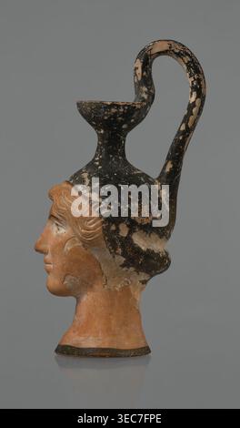 Oinochoe in the shape of a woman's head, Greece, Attica, ca. 450 BC. Oinochoe vessel fashioned from yellowish clay with an orange surface and, in places, shiny black engobe; some elements painted white. The upper part and the headdress are covered with black engobe, on which a white olive wreath stands out. The neck is outlined at the base with a black band. The elongated female face, very carefully modeled, corresponds to the classic Attic head type from the second half of the 5th century. The oval eyes are surrounded by a black outline. The hair is divided by a parting and falls in waves to Stock Photo