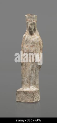 Female terracotta figurine of a standing woman, depicted frontally. Set on a small, hollow, bottomless base, rectangular at the front and oval at the back. A large, rectangular opening at the back shows clear traces of carving in the unfired clay. The woman stands with legs slightly apart, weight on the straight right leg, left leg bent slightly forward. Arms lowered, slightly bent at the elbows; hands hold a cloak at hip level. Head turned slightly to the left on a long neck; elongated face with very faint features; long combed-back hair over the shoulders. A polos on her head is decorated Stock Photo