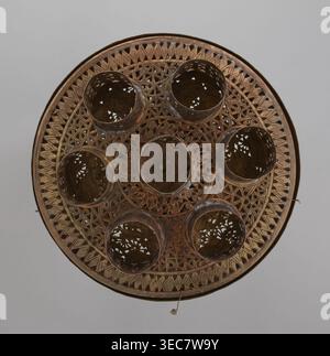 Lamp, 19th–20th century, Damascus, Syria. Metal Islamic lamp with Arabic inscriptions and geometric ornament; cover decorated with a star motif. Disc base with seven holes for glass sleeves glass oil containers missing. Height 40 cm, diameter 32 cm, weight 2.4 kg., A round, ornate object viewed from above with a predominantly brown and bronze color palette, featuring an outer rim and a patterned interior with geometric lattice work and carved scroll-like motifs; a central circular area is surrounded by a group of several hollow cylindrical cups or compartments with perforated sides that reveal Stock Photo