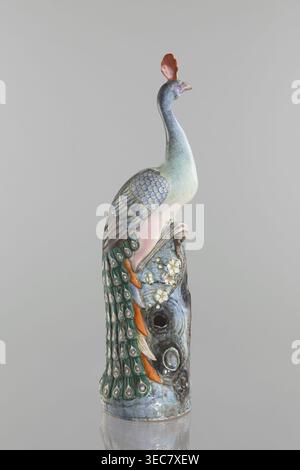 Peacock figurine, Chinese, Qing dynasty 1644–1911, 19th century. Ceramic colored porcelain sculpture depicting a peacock with peacock-feather ornament, images of birds, plum trees, tree trunks, and flowers. Height 54.5 cm; width 14.0 cm; depth 12.5 cm; weight 2.9 kg., A ceramic sculpture of a bird perched on a textured base, shown against a plain pale gray background. The bird has a long curved neck and a small head topped with a crest that is reddish-orange. Its body shows soft gradients of pale green, bluish-gray, and light pink, with layered wing feathers rendered in muted blue-gray tones. Stock Photo