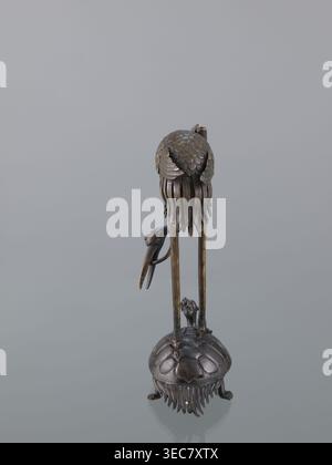Candlestick shokudai, lotuses, turtles, cranes iconography, Japanese art, Japanese culture, Meiji 1868–1912, animals, 2nd half of the 19th century, Japan, candlestick, height 37.0 cm, A tall metallic sculpture with a dark bronze and brass patina stands on a rounded base resembling a shell with textured ridges and fringe-like elements, supported by small clawed feet; two long, slender vertical legs rise from the base to support a rounded upper form covered in overlapping scale-like plates and flowing, ribbon-like strands that hang downward, with a curved beak-like element attached to one side; Stock Photo