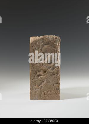 Seal stamp, carved seal showing a dragon with a pearl amid clouds; material: sculpture, Chinese, Qing dynasty 1644–1911, 18th–19th century; dimensions: height 26.5 cm, width 12.2 cm, depth 12.5 cm., A rectangular carved object standing upright on a flat surface, showing a relief design of flowing, sinuous shapes and stylized animal forms on its front face; the material appears to be stone or a stone-like substance with a textured, slightly rough surface and visible cracks and chips; colors visible include shades of beige, tan, light brown, and darker brown accents, set against a neutral Stock Photo