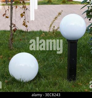 Two white spherical garden lights in a landscaped area. One globe lamp sits on the green grass while the other is mounted on a black post. Paved walkw Stock Photo