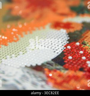 Close-up of a colorful diamond painting with orange, red, and white beads arranged in a floral pattern. Background features a blurred floral design Stock Photo