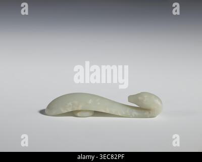 Belt clasp, Chinese, Qing dynasty 1644–1911, 17th–18th century. Milky-white, slightly translucent jadeite; smooth, rounded handle. Full-sculpture carved head with engraved details depicting lizards. Belt buckle; height 2.2 cm, width 10.5 cm, depth 2.1 cm., A smooth pale greenish-white carved object shaped like a stylized animal with a rounded head at one end and a gently curved elongated body, resting on a flat surface with a soft shadow; colors visible include pale green, white, and subtle creamy off-white mottling, set against a neutral light gray to darker gray gradient background. Stock Photo