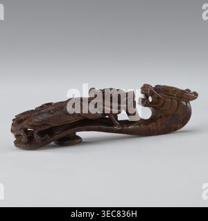 Belt clasp, Chinese; dragons iconography; Qing 1644–1911; 19th century; men's clothing ornament — belts. Shaft: two fighting lizard-dragons. Full-sculpture carved head; openwork shank. Materials/technique: carved. Dimensions: height 3.0 cm, width 12.8 cm, depth 2.8 cm., A carved wooden object in dark brown tones shaped like a stylized creature with an elongated curved body, a pronounced open-mouthed head with visible teeth, and intricately carved flowing details along its back and sides, resting on a light neutral surface against a soft gradient background of pale gray to muted bluish-gray. Stock Photo