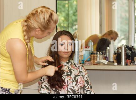 A young woman with long brown hair is getting her hair styled by a blond woman in a beauty salon. Various hair products are visible in the background Stock Photo