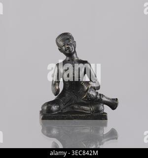 Musician figurine, Southeast Asian art; topic: musicians; culture: Southeast Asia; provenance, 1st half of the 20th century contemporary product; origin: Cambodia; sculpture, figurine; height 6.3 cm, A small dark figurine of a seated person with a glossy black surface and hints of worn gold highlighting, seated cross-legged on a rectangular base with a reflective surface beneath, the figure wearing a fitted upper garment with subtle carved details, one hand resting on the knee and the other holding a rounded object near the lap, the head tilted slightly and showing short textured hair and a Stock Photo