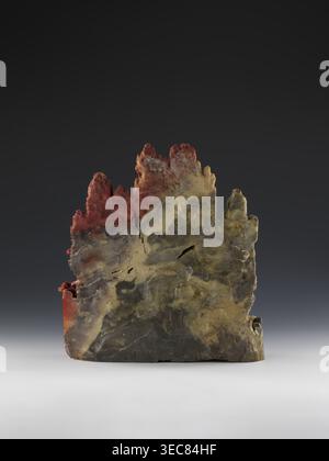 Mountain Landscape with Architecture on a Base - Bas-Relief, Chinese, Qing 1644–1911, mid-19th century. Sculpture depicting mountain landscapes with architectural decorations; height 33.5 cm, width 27.5 cm., A rough, irregular vertical stone slab with jagged peaks and a weathered surface sits on a smooth light-gray base against a dark-gray to nearly black gradient background; the stone shows a mix of colors including muted green, olive, yellowish-beige, dark gray, and areas of rusty red concentrated near the top and along one side, with visible cracks, pitted textures, and layered striations Stock Photo