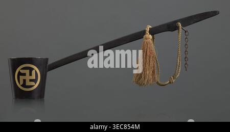 Ritual ladle, Chinese, Qing 1644–1911, Chinese Buddhism, ritual vessel with swastikas painted gold. A chain and silk cord with a tassel hang from the handle. Late 19th century, China. Length 57.5 cm., A long black ladle-like object extends across the image, attached at one end to a short black cup with a circular pale yellow decorative motif on its side, and at the other end to a braided tan cord that holds a tassel of coarse light brown fibers and a short rusty metal chain; the background is a uniform dark gray and the surface beneath the objects reflects them faintly. Stock Photo
