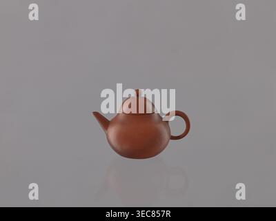 Teapot with lid, Chinese art, Chinese culture, Qing 1644–1911, ceramics, teapots, Yixing style, tea accessories, red stoneware, 19th century, China, Yixing, Jiangsu, vessel, height 6.1 cm, diameter 9 cm, A small round teapot in a reddish-brown clay color sits centered against a smooth light gray background, with a rounded lid topped by a small knob and a curved handle on one side and a short spout on the other, and a soft reflection of the teapot visible below it. Stock Photo