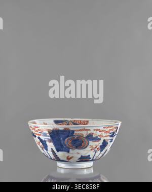 Bowl; maker,  noted. Japanese, Edo period ca. 1680–1720. Colored porcelain vessel, height 12.4 cm, diameter 25.8 cm. Decoration: outside shows a stylized landscape with pagodas; inside features large ornamental rosettes and flowered branches. Subjects: pagodas, plant ornaments, flowers, rosette., A shallow ceramic bowl with a rounded foot, shown against a plain gray background, decorated with a pattern in blue and reddish-orange on a white base; the exterior shows stylized landscape motifs including trees and architectural forms in blue, accented with reddish-orange cloud-like bands and floral Stock Photo