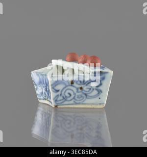 Vessel, ceramics, Chinese art, Chinese, plant ornaments, blue-and-white porcelain, brick-red balls fired from clay, early 20th century, China, vessel; height 3.1 cm, width 7.5 cm, depth 4.6 cm, A small ceramic object sits on a reflective surface against a gray background, shaped like a shallow rectangular boat with rounded inner dividers and a flat top that holds a group of three rounded beads. The ceramic has a white base color with blue painted swirling floral patterns on its sides and faint brown spots in places, and the beads on top are a muted reddish-pink. The surface reflects the object Stock Photo