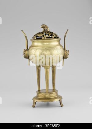 Censer with a cover, Censer with two long handles with mascarons. Decoration: two reserves — fruits and leafy twigs, animals and birds among trees. Early 20th century, Korea. Vessel censer. Height 15.0 cm; diameter 9.8 cm., A brass vessel with a rounded body stands on a raised circular base and a group of tall tripod legs; the body has incised decorative leaf and vine patterns and a warm golden color with areas of darker patina. Two curved vertical handles extend upward from opposite sides, each attached to a sculpted animal head in a similar golden tone. The lid is pierced with an openwork Stock Photo