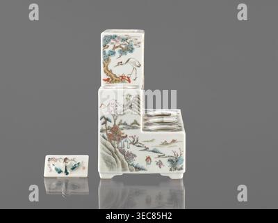Pedestal for a writing set three-stage, late 19th century, China. Colored porcelain with raised central part; height 18.9 cm, width 17.9 cm, depth 11.0 cm. Decoration: ceramics with flowers, figural scenes, butterflies, landscape motifs in Qing style Qing, 1644–1911. Function: writing accessories., A small ceramic stepped object and a separate rectangular ceramic lid sit on a reflective surface against a plain gray background; the stepped piece is primarily white with painted scenes in colors including blue, green, brown, red, pink, and black, showing landscape elements like trees, mountains, Stock Photo