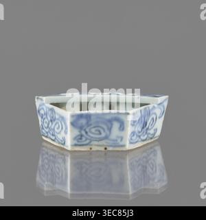 Vessel, gift, repossession Krakow, ceramics, Chinese art, Chinese culture, plant ornaments, blue-and-white porcelain, brick-red balls fired from clay, early 20th century, China, vessel, height 2.6 cm, width 7.4 cm, depth 4.7 cm, A small hexagonal ceramic dish with scalloped top edges, glazed in pale white and decorated with blue painted swirling floral motifs on its sides, sitting on a reflective surface that mirrors the dish; visible colors include white, blue, gray, and a few small brown spots on the base. Stock Photo