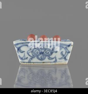 Vessel, ceramic, blue-and-white porcelain with plant ornaments and brick-red fired clay balls, early 20th century, China; height 3.1 cm, width 7.5 cm, depth 4.6 cm., A small rectangular ceramic object with sloping sides sits centered against a solid grey background, decorated with blue swirling floral-like brushwork on a white base; three round reddish-brown spheres rest in shallow depressions along the top edge, and the object and its decorative pattern are reflected faintly on the glossy surface beneath. Stock Photo