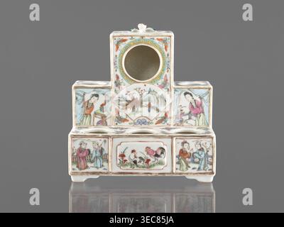 Pedestal for a writing set three-stage, colored porcelain, late 19th century, China; Qing style Qing, 1644–1911. Decorated with ceramics flowers, butterflies, landscape motifs and figural scenes; raised central part. Function: writing accessories. Dimensions: height 18.9 cm; width 17.9 cm; depth 11.0 cm., A small ceramic object with a stepped rectangular form sits against a gray background, predominantly white with painted decoration in colors including pink, green, blue, yellow, orange, brown, and black. The piece has a central circular opening near the top and a few round openings along the Stock Photo