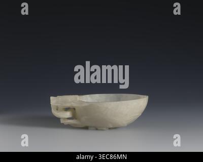 A vessel for rinsing brushes, Chinese, Qing dynasty 1644–1911, 19th century. Painted decoration of peaches iconography; painting implements. Vessel. Height 3.5 cm; width 11.0 cm; depth 7.4 cm., A smooth pale bowl with a creamy off-white color sits on a flat surface against a dark gray to black gradient background, showing a carved handle-like decorative element on one side and subtle translucent veining throughout the stone. Stock Photo